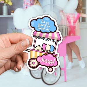 May include: A pink and yellow cotton candy cart with a blue cloud above it that says "Tis so sweet to trust in Jesus!" The cart has a pink and white striped awning and is on wheels.