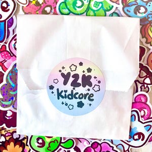 May include: A white paper bag sealed with a round sticker. The sticker has a pastel rainbow gradient background with the words "Y2K Kidcore" in black and white. The bag is surrounded by colorful stickers.