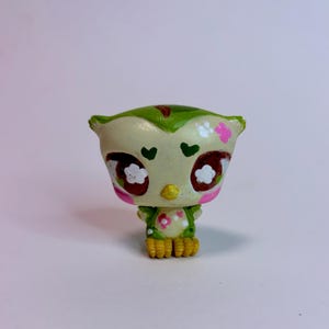 May include: A small owl figurine with a cream-colored head and green body. The owl features large brown eyes with white flower accents, green heart-shaped markings above the eyes, pink cheek spots, and yellow feet. The body has pink flower details.