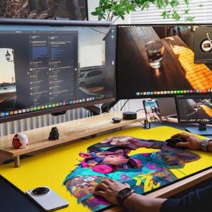 May include: A large rectangular desk mat featuring a vibrant yellow background with a colourful graphic illustration of a monkey wearing headphones and a cap. The mat is positioned on a wooden desk surface beneath two computer monitors.