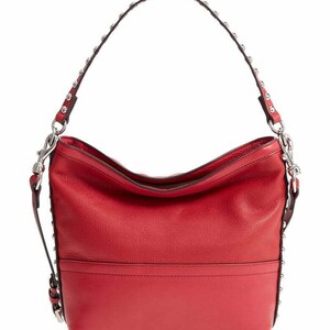 Op de afbeelding: Een felrode hobo-tas met een getextureerd oppervlak en een glad horizontaal paneel aan de onderkant. De tas heeft een enkele schouderband versierd met zilverkleurige metalen studs over de lengte, verbonden aan de tas met zilverkleurige hardware.