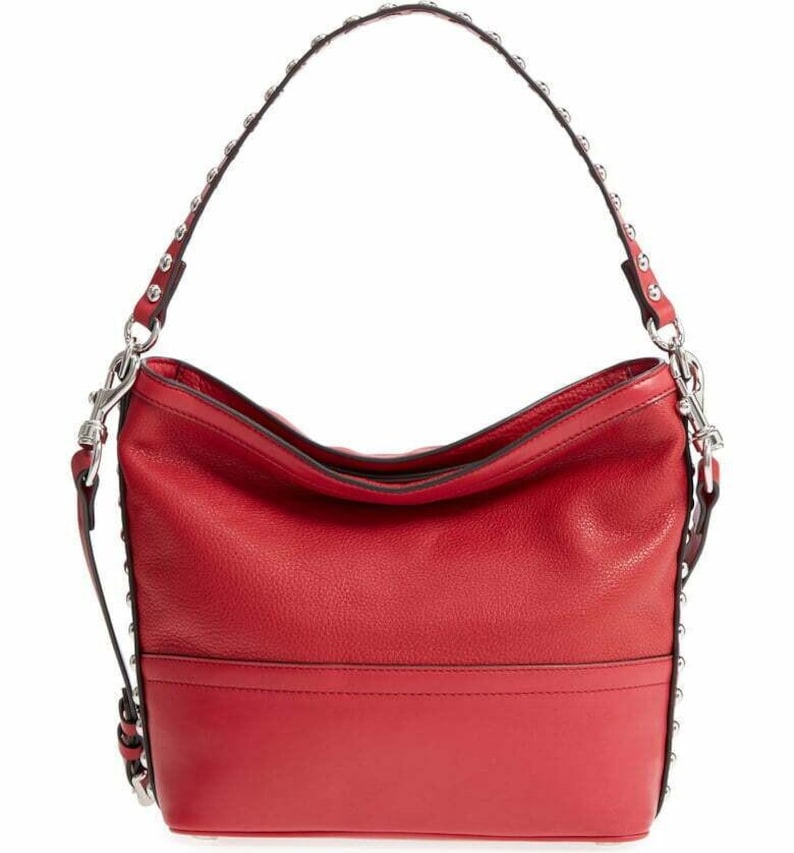 Op de afbeelding: Een felrode hobo-tas met een getextureerd oppervlak en een glad horizontaal paneel aan de onderkant. De tas heeft een enkele schouderband versierd met zilverkleurige metalen studs over de lengte, verbonden aan de tas met zilverkleurige hardware.