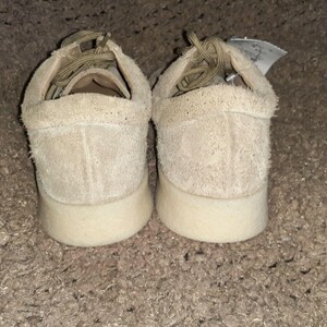 May include: A pair of beige suede low-top sneakers featuring a textured, fuzzy exterior, matching beige laces, and a thick, smooth off-white rubber sole. A small white tag is attached to the heel area of the right shoe, visible from the back.