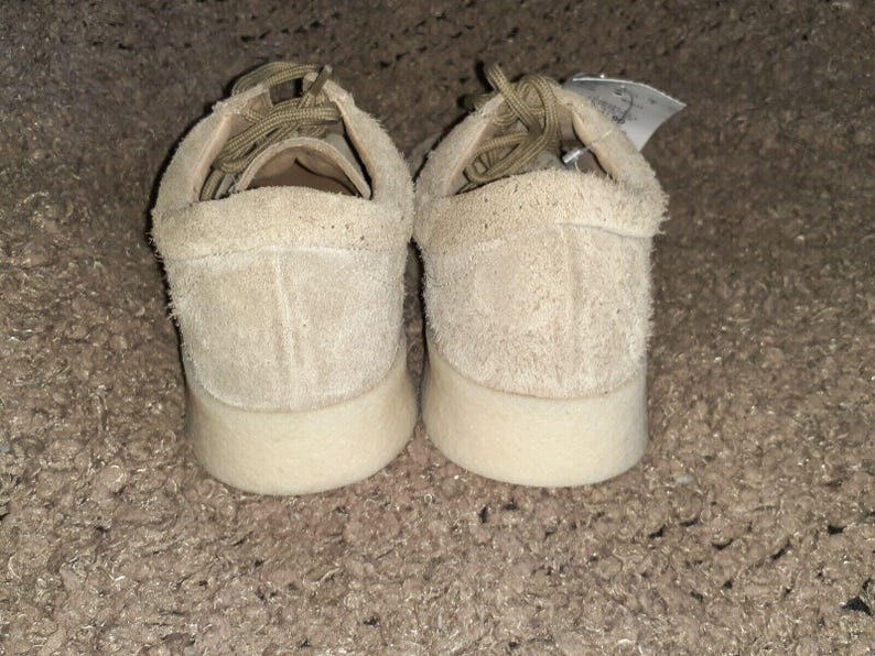 May include: A pair of beige suede low-top sneakers featuring a textured, fuzzy exterior, matching beige laces, and a thick, smooth off-white rubber sole. A small white tag is attached to the heel area of the right shoe, visible from the back.
