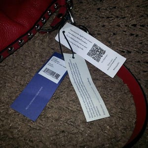 Op de afbeelding: Een rode leren handtas met zilverkleurige metalen studs langs de rand. Aan de tas hangen drie rechthoekige productlabels, waaronder een met een QR-code, een met stijlinformatie en een met onderhoudsinstructies voor het leer.