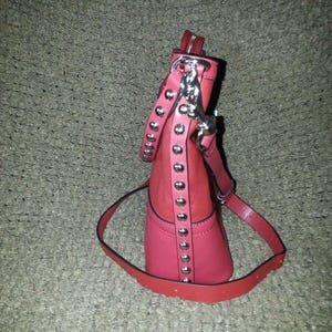 Op de afbeelding: Een tweekleurige rode en roze handtas met een verticale strook zilverkleurige metalen studs aan de zijkant. De tas is voorzien van een bijpassende rode schouderriem bevestigd met zilverkleurige hardware en een ritssluiting aan de bovenzijde.