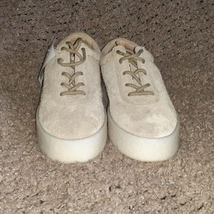 May include: A pair of beige suede lace-up sneakers featuring thick, white platform soles. The shoes have matching beige laces and a rounded toe design, with a small white tag attached to the left shoe near the eyelets, presenting a clean look.