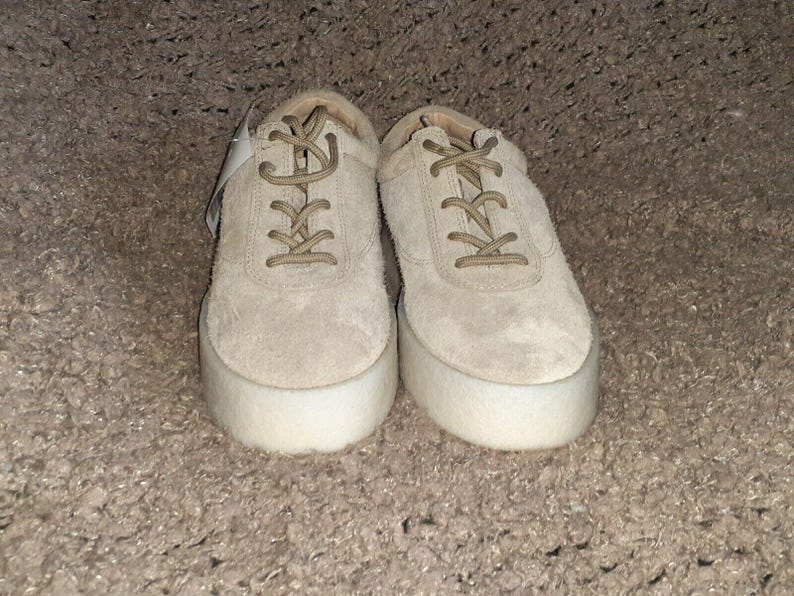 May include: A pair of beige suede lace-up sneakers featuring thick, white platform soles. The shoes have matching beige laces and a rounded toe design, with a small white tag attached to the left shoe near the eyelets, presenting a clean look.
