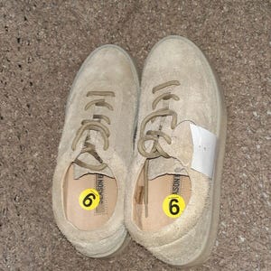 May include: A pair of beige suede sneakers with matching laces and a low-top silhouette. Each shoe features a yellow circular sticker with the number 6 inside the footbed, and one shoe has a white rectangular tag attached to the laces.