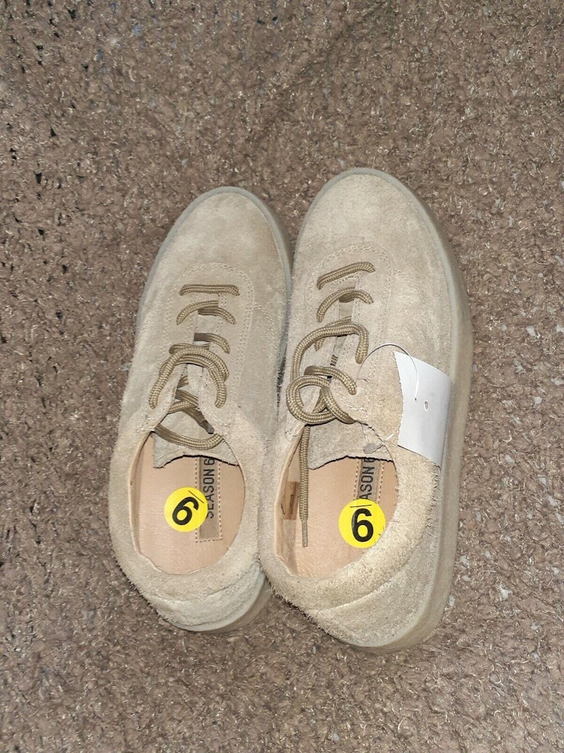 May include: A pair of beige suede sneakers with matching laces and a low-top silhouette. Each shoe features a yellow circular sticker with the number 6 inside the footbed, and one shoe has a white rectangular tag attached to the laces.