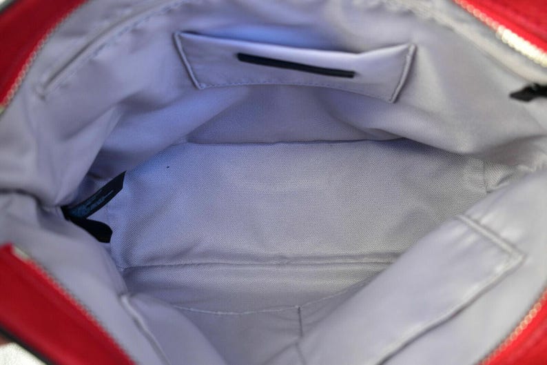 Op de afbeelding: De binnenkant van een rode handtas met een lichtgrijze stoffen voering. De tas bevat een rechthoekig steekvak op de achterwand met een zwarte rand en een klein zwart stoffen label bevestigd aan de zijnaad bij de bovenste opening.