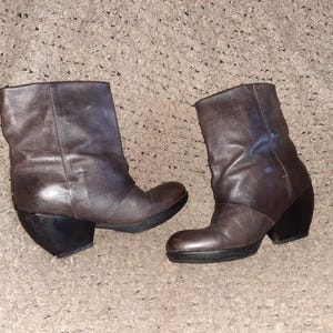 May include: A pair of dark brown leather ankle boots featuring a rounded toe, a mid-calf shaft with a slightly slouchy appearance, and a thick, solid black block heel with a matching black sole that extends slightly under the front of the boot.