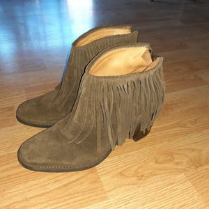 May include: A pair of olive green suede ankle boots featuring a rounded toe, a stacked heel, and a decorative fringe layer around the collar. The boots have a zipper closure at the back and a tan interior lining, showcasing a textured finish.