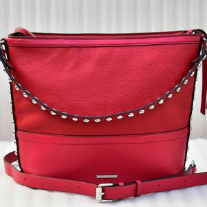 Op de afbeelding: Een felrode hobo-schoudertas met een gestructureerd bovenpaneel en een glad onderpaneel. De tas is voorzien van een gebogen band met zilverkleurige metalen studs aan de voorzijde en heeft een verstelbare schouderriem.