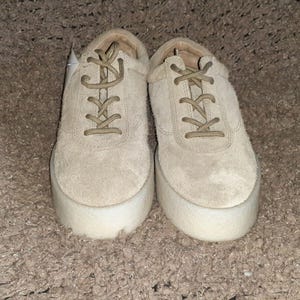 May include: A pair of beige suede sneakers featuring a lace-up closure with matching beige laces and a thick, white, textured platform sole. The shoes have a rounded toe design and a low-top silhouette, showcasing a uniform light tan color throughout.