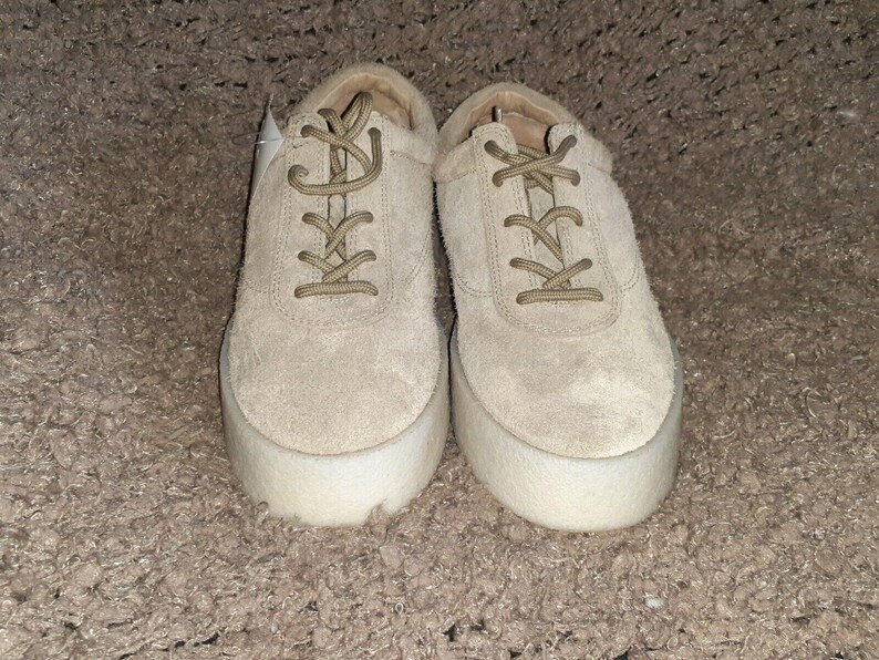 May include: A pair of beige suede sneakers featuring a lace-up closure with matching beige laces and a thick, white, textured platform sole. The shoes have a rounded toe design and a low-top silhouette, showcasing a uniform light tan color throughout.