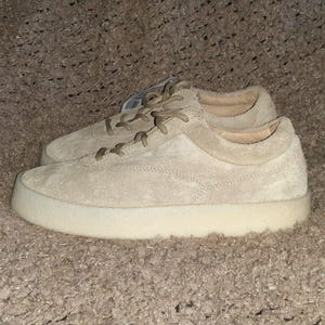 May include: A pair of low-top sneakers in a light beige suede material. The shoes feature matching beige laces, a rounded toe, and a thick, solid off-white rubber sole. A small white tag is visible near the eyelets of the top shoe.