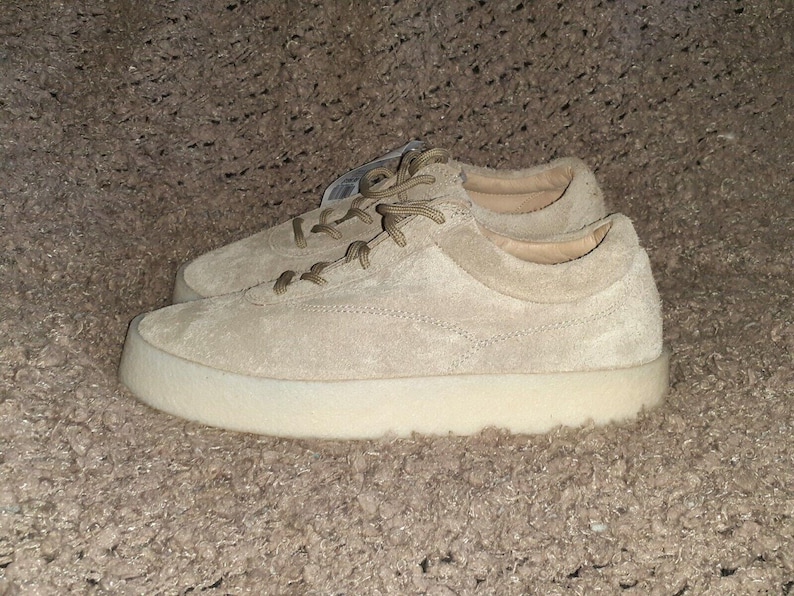May include: A pair of low-top sneakers in a light beige suede material. The shoes feature matching beige laces, a rounded toe, and a thick, solid off-white rubber sole. A small white tag is visible near the eyelets of the top shoe.