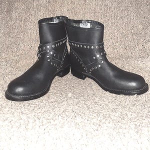 May include: A pair of black leather ankle boots featuring a rounded toe, a low block heel, and a decorative strap across the mid-foot adorned with silver-toned metal studs that wrap around the ankle and side of the boot shaft for a detailed finish.
