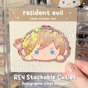 May include: A Resident Evil tsum sticker set featuring a holographic vinyl sticker of a stylized character with brown hair and blue eyes. The packaging includes the text resident evil tsum sticker set and pixelclaire. The sticker has a shiny finish.