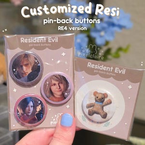 May include: Two clear plastic packages containing pin-back buttons. The left package features three buttons with character portraits labeled Resident Evil. The right package contains one button with a brown teddy bear graphic labeled Resident Evil.