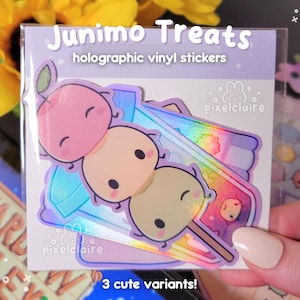 Könnte beinhalten: Eine durchsichtige Kunststoffverpackung mit holografischen Vinylaufklebern mit der Aufschrift Junimo Treats. Die Aufkleber zeigen drei gestapelte, runde Cartoon-Figuren in Rosa, Creme und Hellgrün auf einem Stiel, mit dem Text pixelclaire.