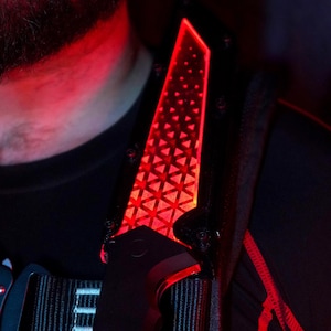 May include: A black tactical vest with a red light-up panel on the shoulder strap. The vest has a black strap with a silver buckle and a black and white logo that says "Nemo's Forge".