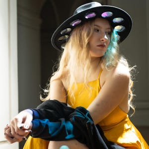 May include: A woman wearing a yellow dress and a black hat with circular cutouts. The hat has iridescent accents. She is holding a black jacket in front of her.