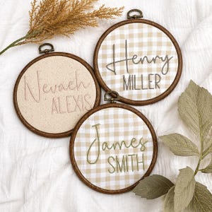 May include: Three circular wooden embroidery hoops containing fabric with embroidered names. One hoop features the name Nevaeh Alexis on cream fabric, while the others display Henry Miller and James Smith on beige and white gingham fabric.