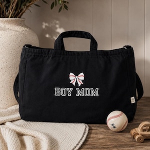 May include: Black tote bag with the embroidered text "BOY MOM" and a bow design. A baseball sits next to the bag. The bag has two handles and a shoulder strap. The bag is on a wooden surface.