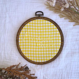 May include: Round wooden embroidery hoop with a yellow and white gingham fabric. The name "Amelia Grace" is embroidered in yellow thread across the fabric. The hoop has a dark brown finish and a metal clasp.