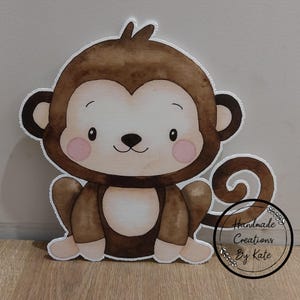 May include: A brown and white cartoon monkey cutout with a curled tail. The monkey has a cute, friendly face with pink cheeks and a small black nose. The cutout is made of cardstock and is perfect for a child's birthday party or nursery decor.