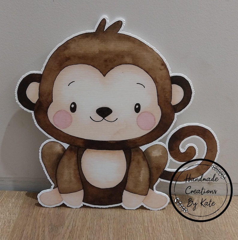 May include: A brown and white cartoon monkey cutout with a curled tail. The monkey has a cute, friendly face with pink cheeks and a small black nose. The cutout is made of cardstock and is perfect for a child's birthday party or nursery decor.