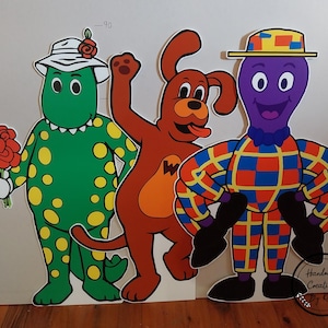 May include: Four colourful cardboard cutouts of characters from the children's television show 'The Wiggles'. The characters are a green dinosaur wearing a pink hat, a red dog, a purple octopus wearing a yellow and red hat, and a yellow and red striped character with a purple head.