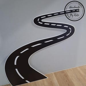 May include: A black and white road-shaped wall decal with a winding design. The road has white dashed lines and a white border. The decal is placed on a gray wall and the text "Handmade Creations By Kate" is in a circle.
