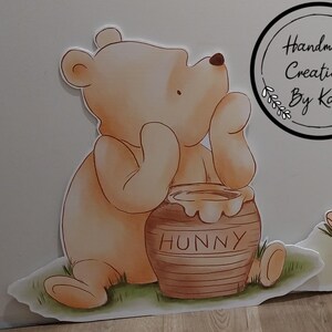 Puede incluir: Un personaje de dibujos animados de Winnie de Pooh sosteniendo una olla de miel que dice "Hunny" en la parte delantera. El personaje est&aacute; sentado en un parche de hierba verde.