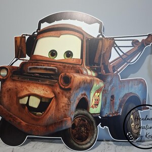 May include: A large, cardboard cutout of the character Mater from the animated film Cars. The cutout features Mater's rusty brown and blue body, large eyes, and a friendly grin. The image includes a tow hook and the number 95.