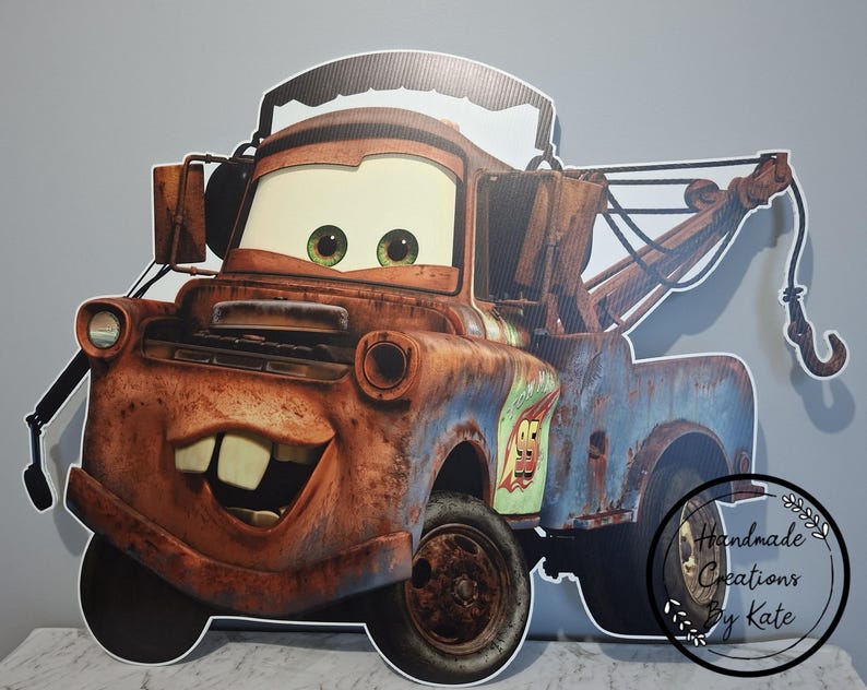 May include: A large, cardboard cutout of the character Mater from the animated film Cars. The cutout features Mater's rusty brown and blue body, large eyes, and a friendly grin. The image includes a tow hook and the number 95.