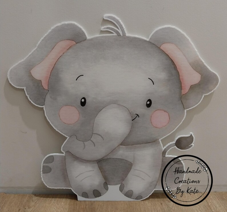 May include: A grey and pink elephant cutout with a watercolour design. The elephant is sitting with its trunk in its mouth. The cutout has a white background and a small white circle with the text "Handmade Creations By Kate".