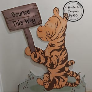 Puede incluir: Un recorte de madera de Tigger de Winnie the Pooh sosteniendo un cartel que dice "Bounce This Way". El recorte est&aacute; pintado de naranja y negro con un fondo de hierba verde.