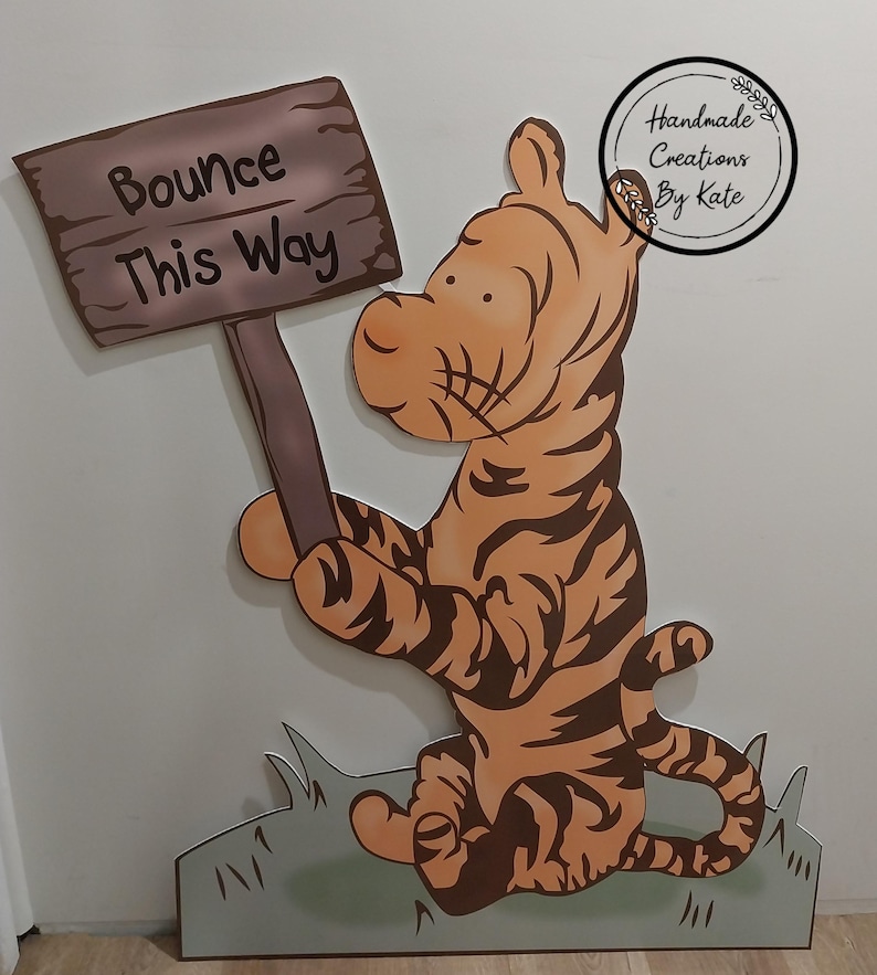 Puede incluir: Un recorte de madera de Tigger de Winnie the Pooh sosteniendo un cartel que dice "Bounce This Way". El recorte est&aacute; pintado de naranja y negro con un fondo de hierba verde.