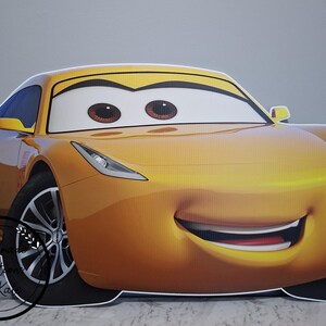 May include: A large, smiling, yellow cartoon car with big brown eyes and a red mouth. The car has black tires and a yellow spoiler. The car is a cutout, likely for a party or event.