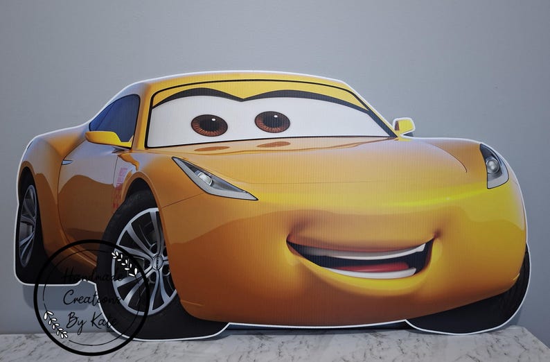 May include: A large, smiling, yellow cartoon car with big brown eyes and a red mouth. The car has black tires and a yellow spoiler. The car is a cutout, likely for a party or event.