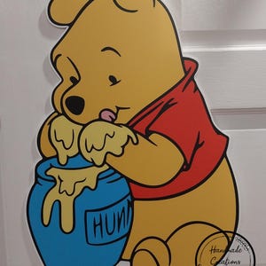 Puede incluir: Un oso de dibujos animados amarillo que lleva una camisa roja est&aacute; sosteniendo un tarro de miel azul con la palabra "HONEY" escrita en &eacute;l. El oso est&aacute; lamiendo la miel que gotea del tarro.