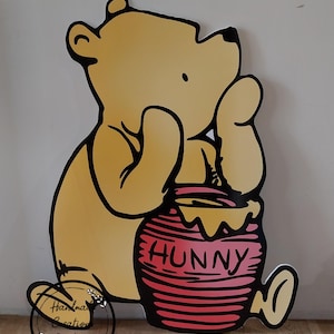 Puede incluir: Un oso de dibujos animados amarillo sosteniendo un tarro de miel rojo con la palabra "Hunny" impresa en &eacute;l. El oso est&aacute; de pie sobre una superficie de madera.