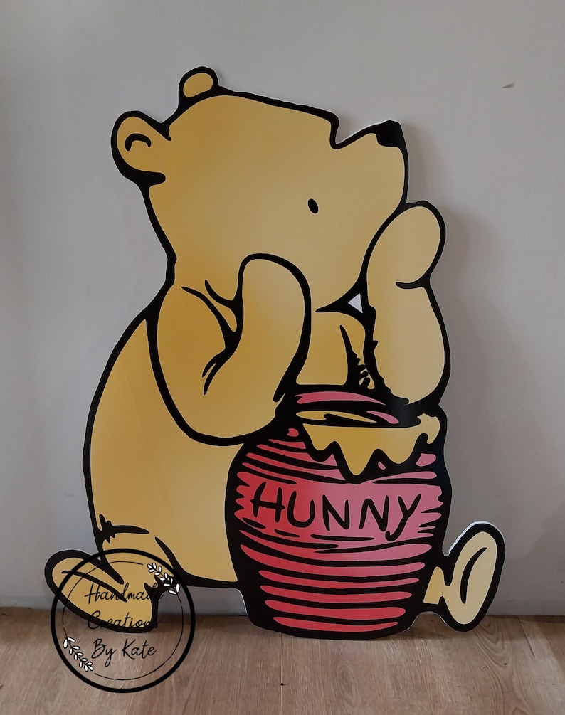 Puede incluir: Un oso de dibujos animados amarillo sosteniendo un tarro de miel rojo con la palabra "Hunny" impresa en &eacute;l. El oso est&aacute; de pie sobre una superficie de madera.