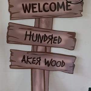 Puede incluir: Un letrero de madera con tres tablas, cada una con una inscripci&oacute;n diferente. La tabla superior dice "WELCOME", la tabla del medio dice "HUNDRED" y la tabla inferior dice "AKER WOOD". El letrero est&aacute; pintado en tonos marrones y beige.
