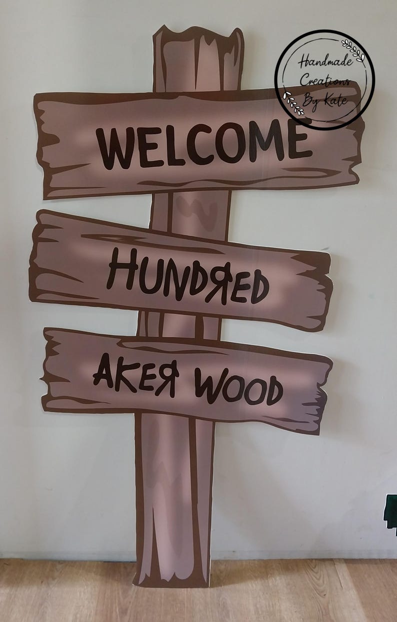 Puede incluir: Un letrero de madera con tres tablas, cada una con una inscripci&oacute;n diferente. La tabla superior dice "WELCOME", la tabla del medio dice "HUNDRED" y la tabla inferior dice "AKER WOOD". El letrero est&aacute; pintado en tonos marrones y beige.