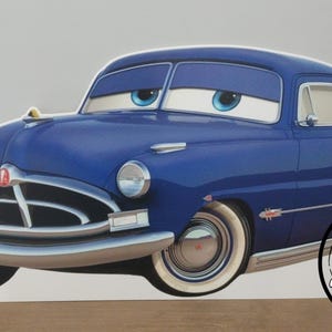 May include: A blue cartoon car with large eyes and a smiling expression. The car has white-walled tires and chrome accents. The car is a vintage model, likely inspired by the Hudson Hornet. The image includes the text "Handmade Creations By Kate".