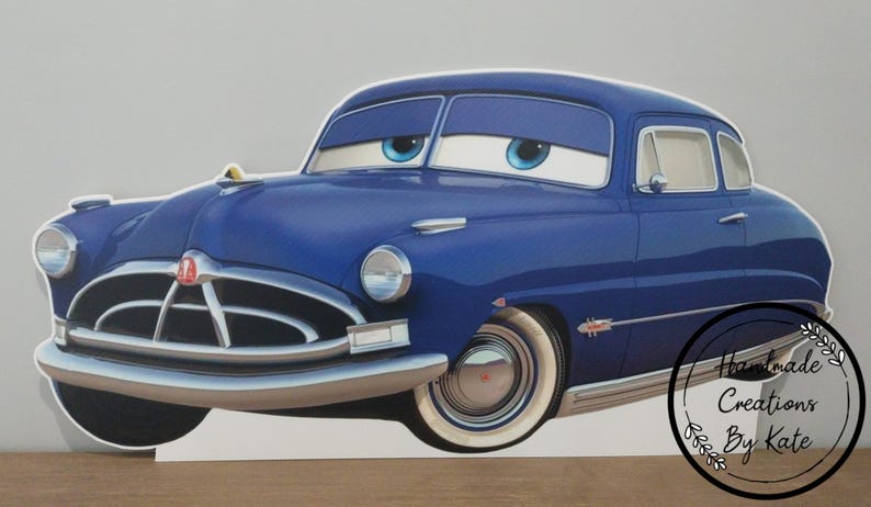 May include: A blue cartoon car with large eyes and a smiling expression. The car has white-walled tires and chrome accents. The car is a vintage model, likely inspired by the Hudson Hornet. The image includes the text "Handmade Creations By Kate".